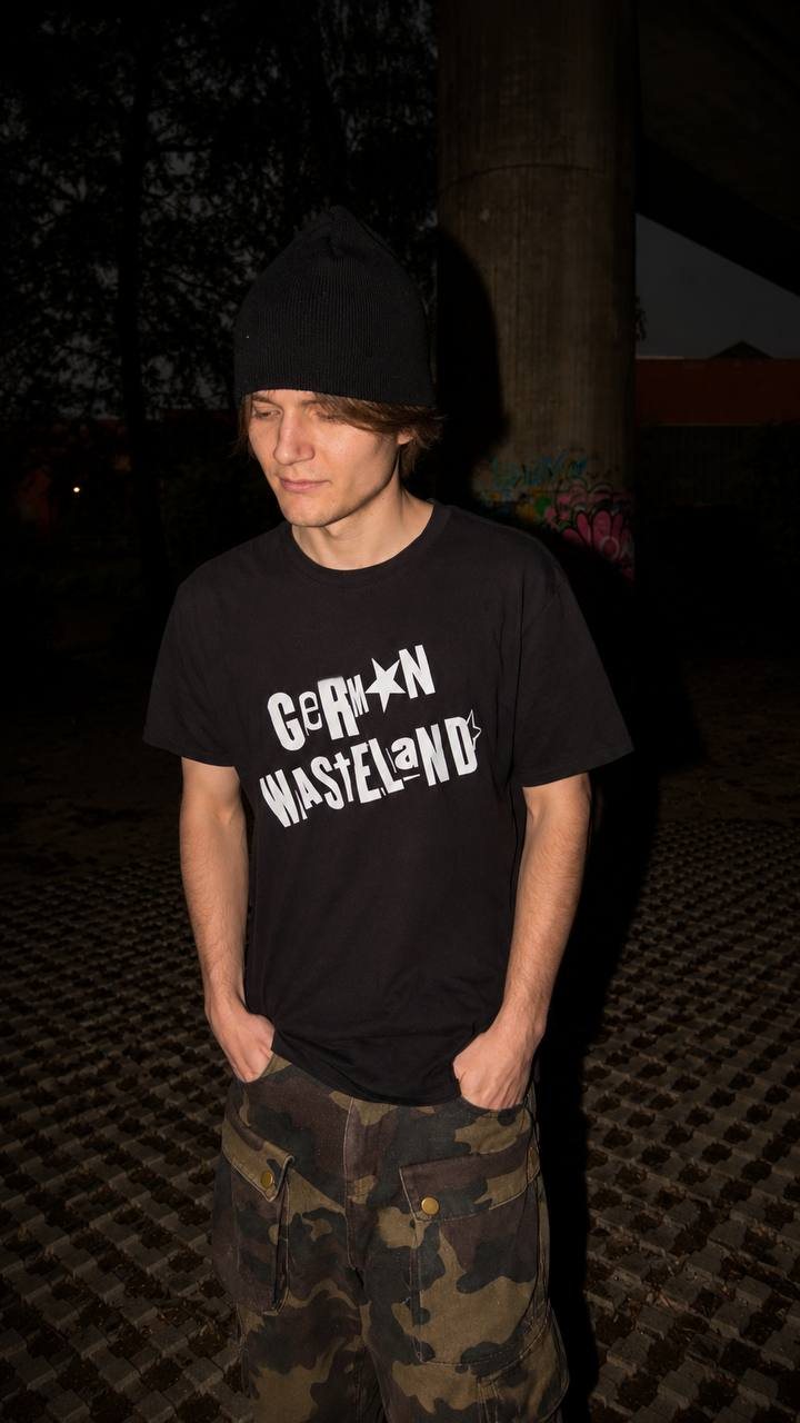 German Wasteland black graphic T-shirt editorial photo with beanie, camouflage cargo pants and nighttime concrete underpass background