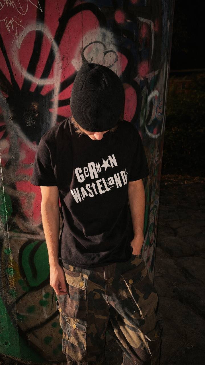 German Wasteland black logo T-shirt close editorial photo with beanie, camouflage cargo pants and graffiti wall background